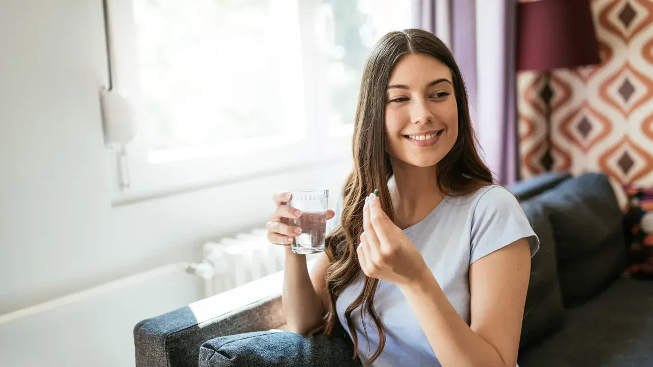 Smiling young woman holding a green supplement capsule and a glass of water, illustrating how to take Alpha GPC supplements