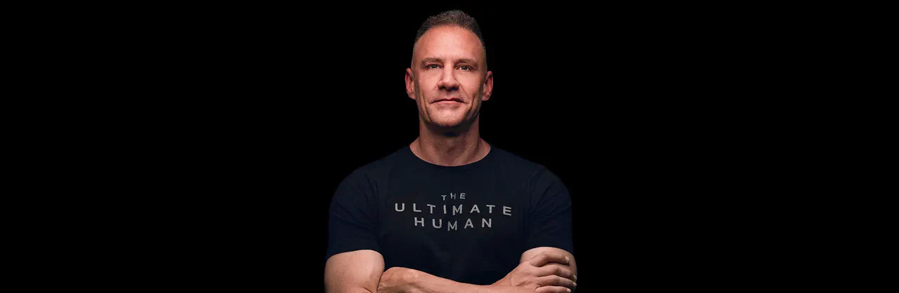 Gary Brecka in a black The Ultimate Human t-shirt standing with arms crossed against a dark background