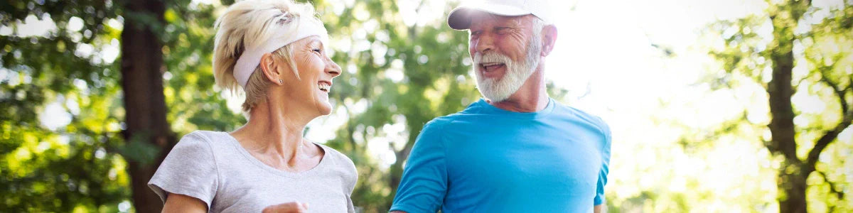 Two older adults – a woman and a man – smiling and jogging together through a sunlit green leafy park in athletic wear