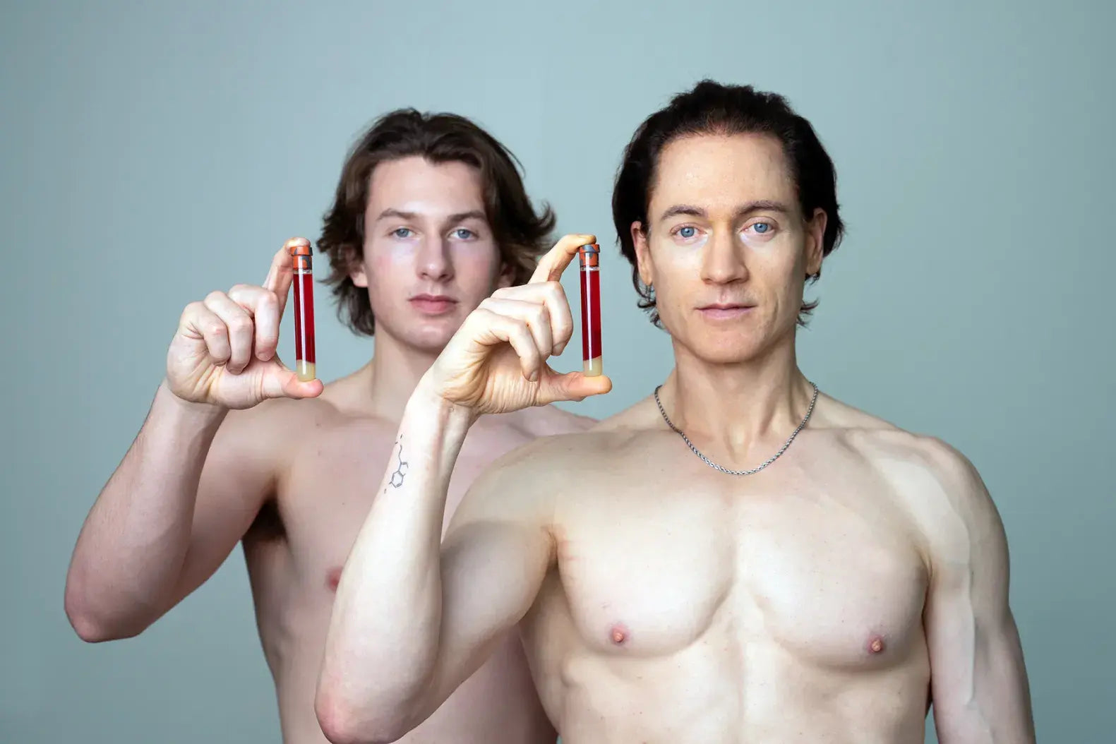 Bryan Johnson and his son each holding up a small vial of blood against a grey-green studio background, depicting their plasma exchange experiment