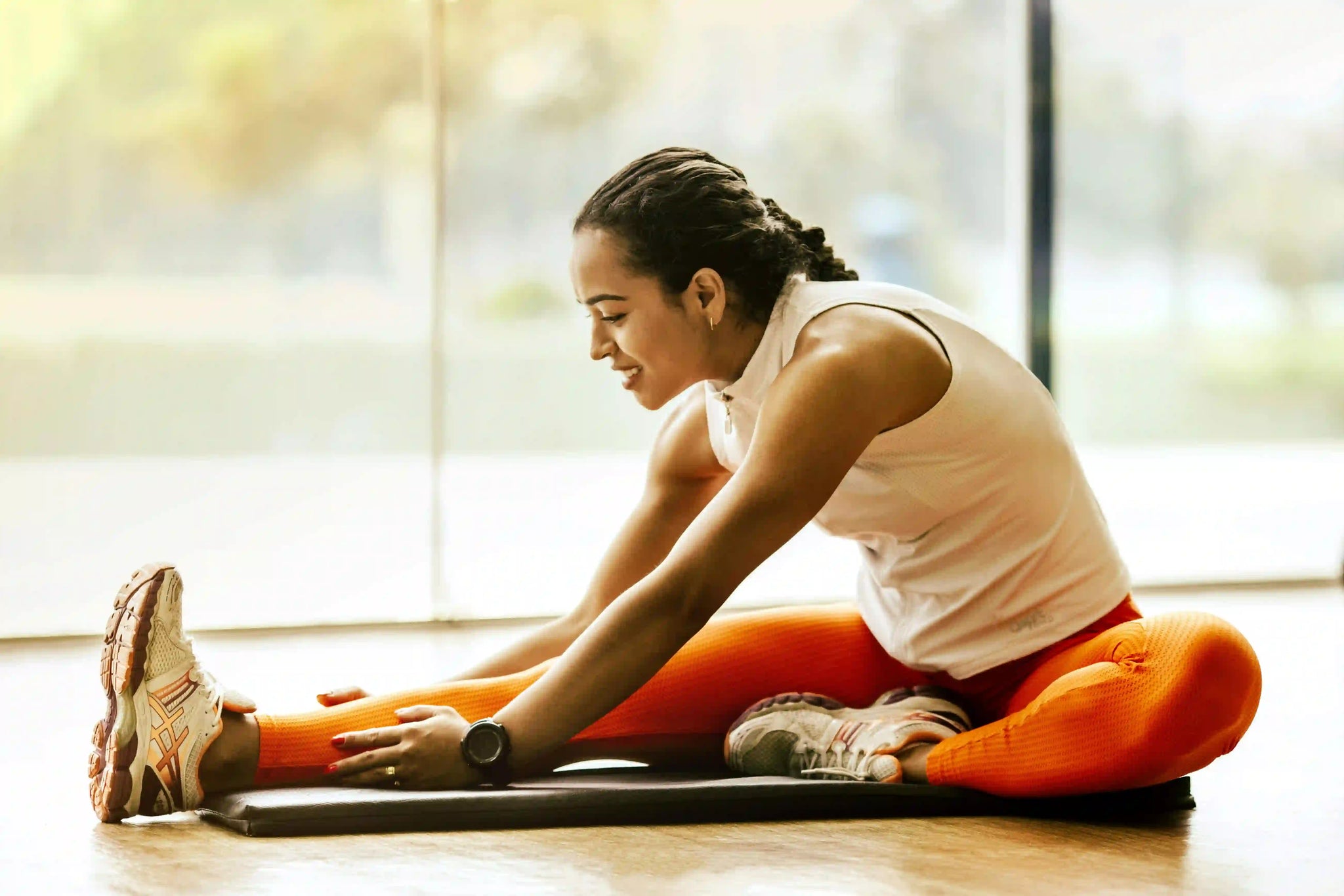 Smiling woman in white tank top and orange leggings doing a seated hamstring stretch on a gym mat, with large floor-to-ceiling windows behind her