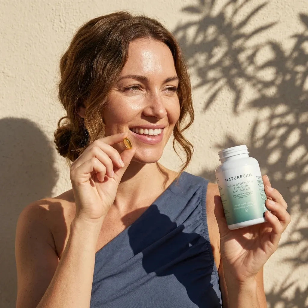 Naturecan High Strength Omega-3 supplement lifestyle – smiling woman holding bottle and an amber softgel capsule in sunlight against cream wall with leaf shadows