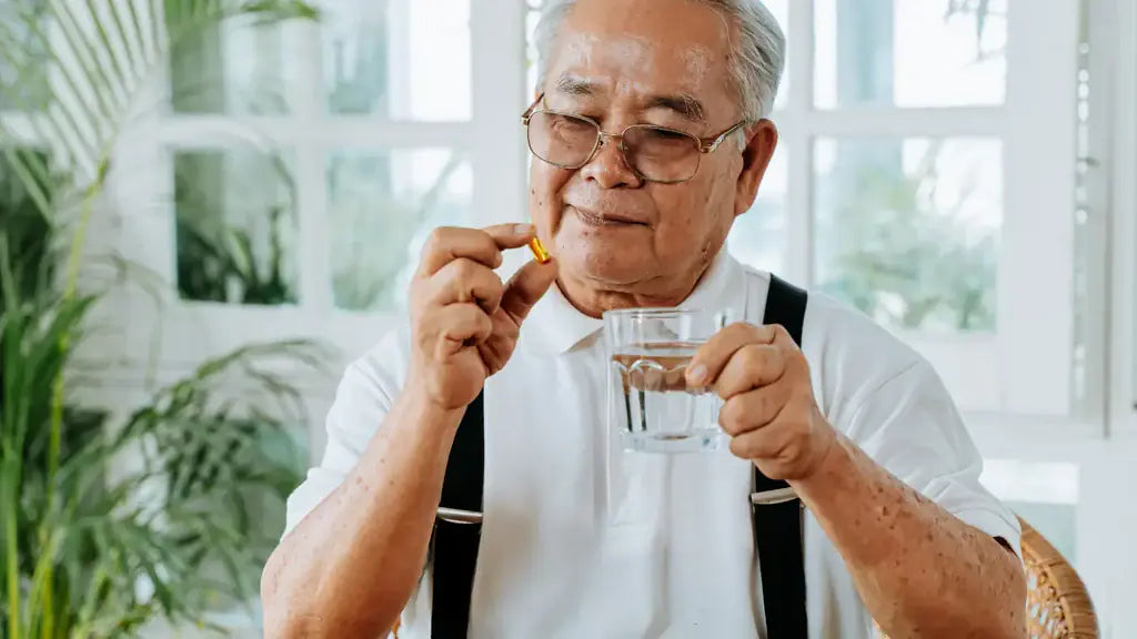 Elderly Asian man with glasses holding a yellow supplement capsule and a glass of water, illustrating AKG supplementation for longevity