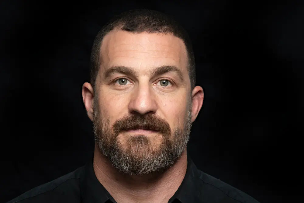 Close-up portrait of Andrew Huberman, neuroscientist and podcast host, with short dark hair and beard, wearing a black shirt against a dark background