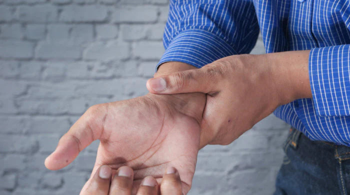 Man in a blue striped shirt gripping his wrist with his other hand against a white brick wall background, indicating joint pain and arthritis symptoms