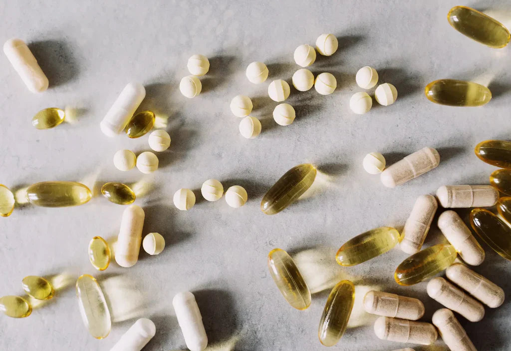 Assortment of supplement pills, capsules, and tablets in various shapes and colours including white, yellow, and beige scattered across a grey marble surface
