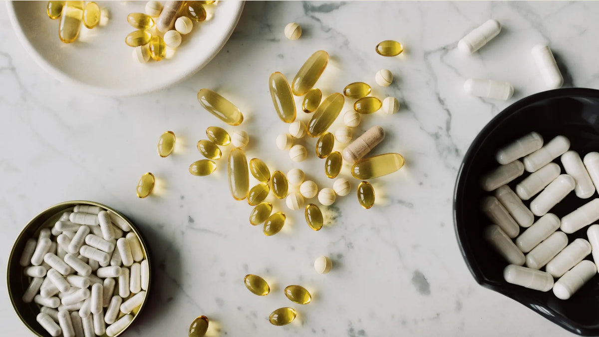 Top-down view of various supplement capsules and softgels – golden softgels, white capsules, and beige tablets – scattered on a marble surface with small bowls