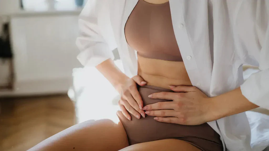 Woman in a white shirt sitting and holding her lower abdomen, depicting discomfort associated with bloating