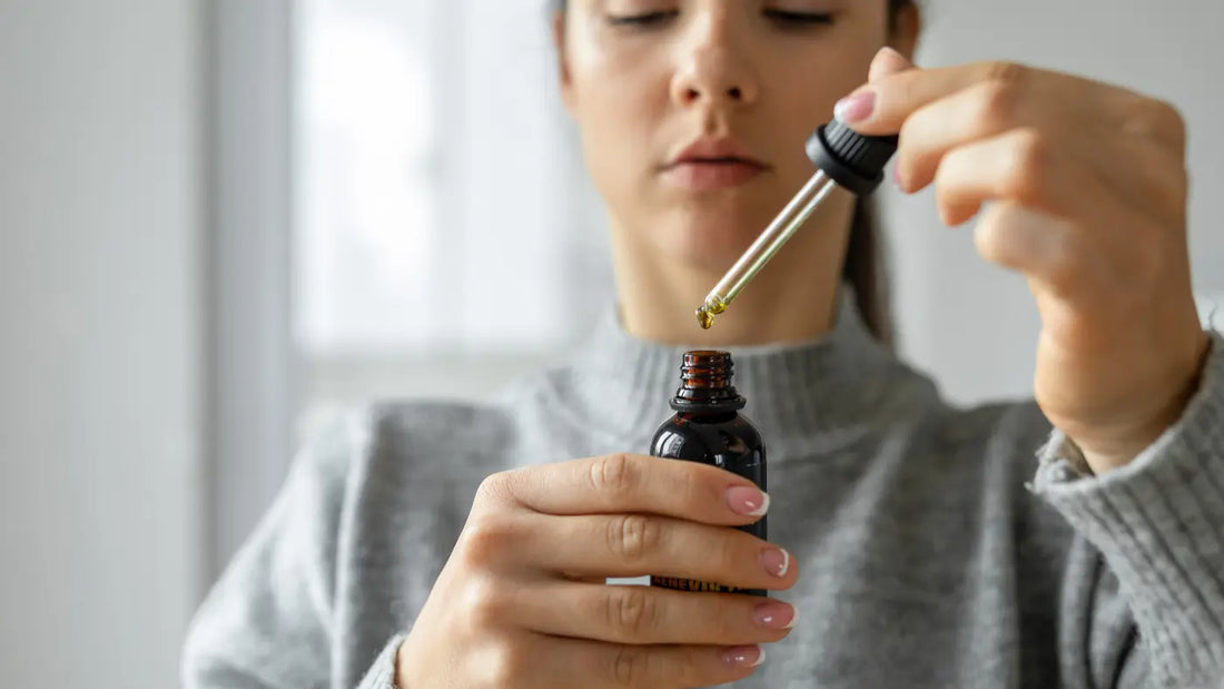 Woman in a grey knit sweater holding a small dark amber CBD oil dropper bottle and carefully lifting the dropper to examine a drop of oil