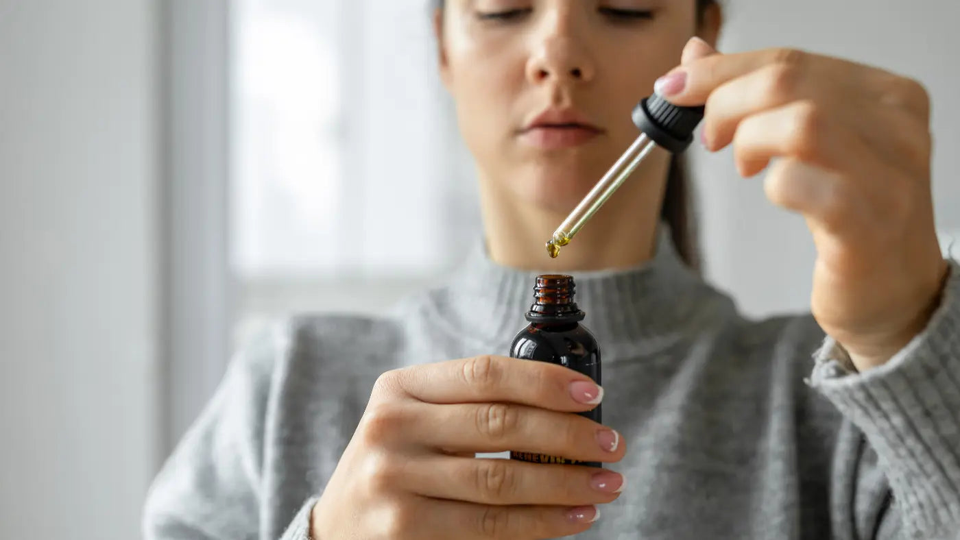 Woman in a grey knit sweater holding a small dark amber CBD oil dropper bottle and carefully lifting the dropper to examine a drop of oil