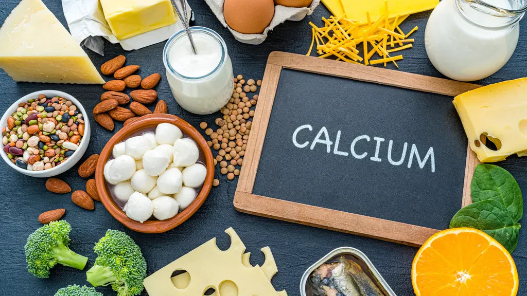 Chalk blackboard with CALCIUM written in white text surrounded by calcium-rich foods including dairy products, eggs, broccoli, almonds, sardines, cheese, and an orange slice