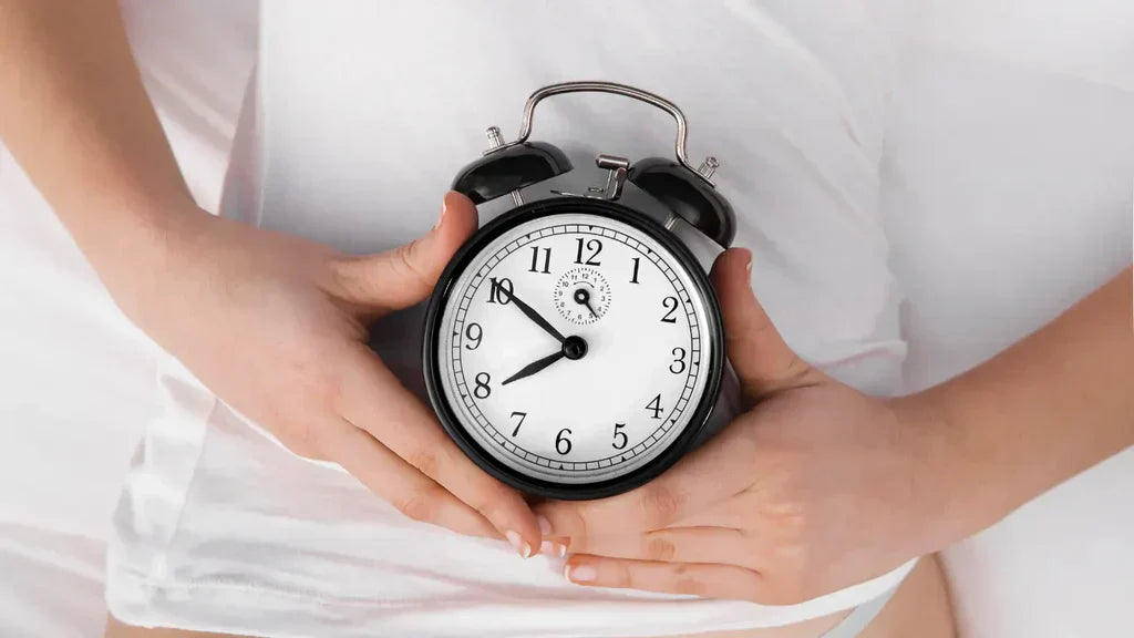 Person holding a classic black alarm clock against their white-clothed abdomen, symbolising the concept of biological versus chronological age
