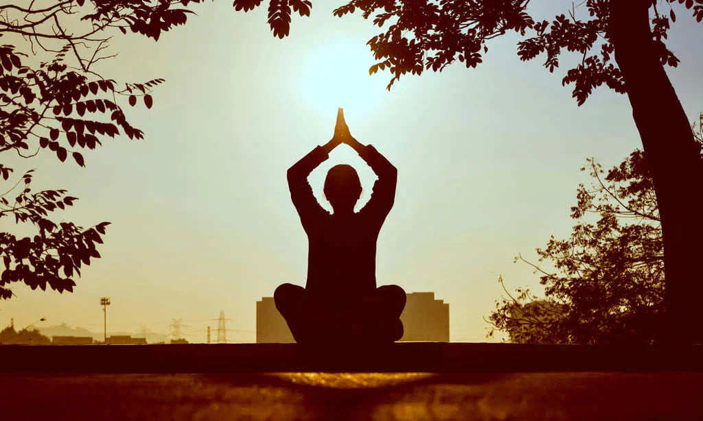 Silhouette of a person sitting in a yoga lotus pose with hands raised overhead, set against a warm golden sunset sky framed by trees