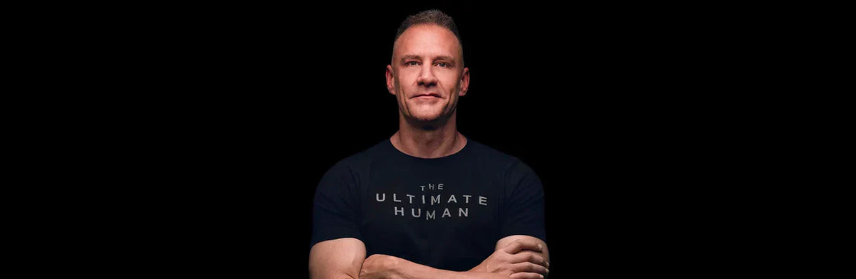 Gary Brecka in a black The Ultimate Human t-shirt standing with arms crossed against a dark background
