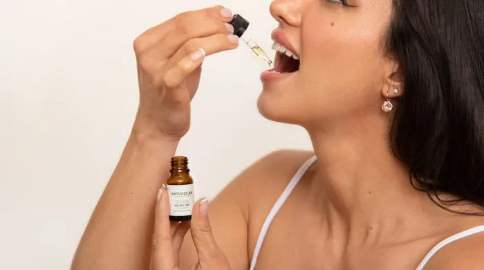 Woman placing Naturecan CBD oil drops under her tongue using a dropper, holding the amber oil bottle in her other hand against a cream background