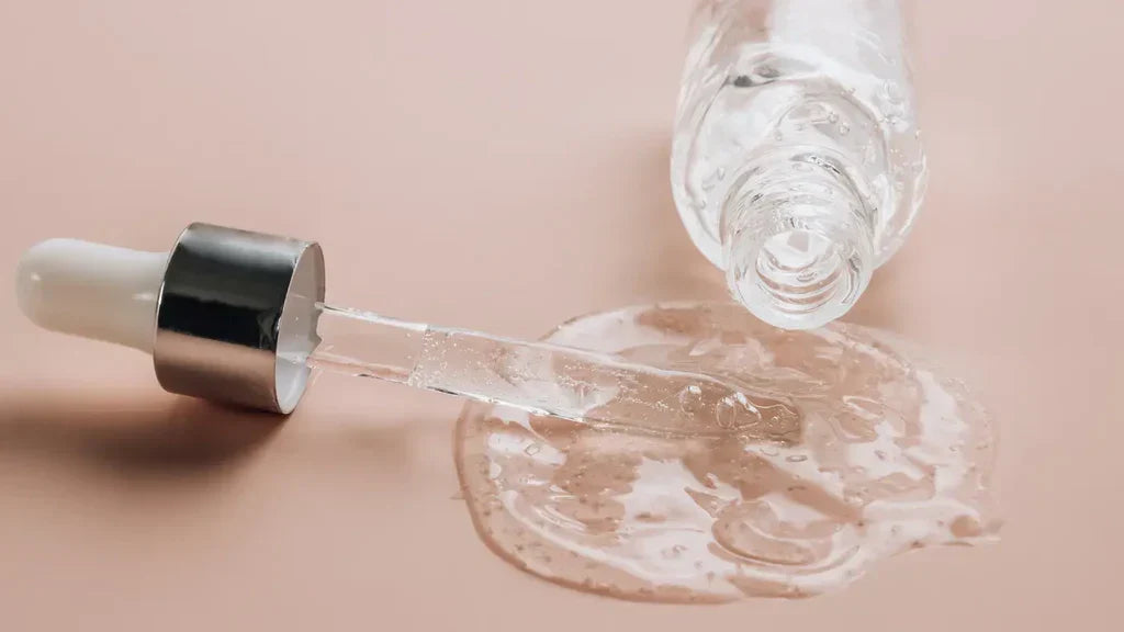 Glass dropper bottle of hyaluronic acid serum on its side on a pink background, with clear gel spread across the surface