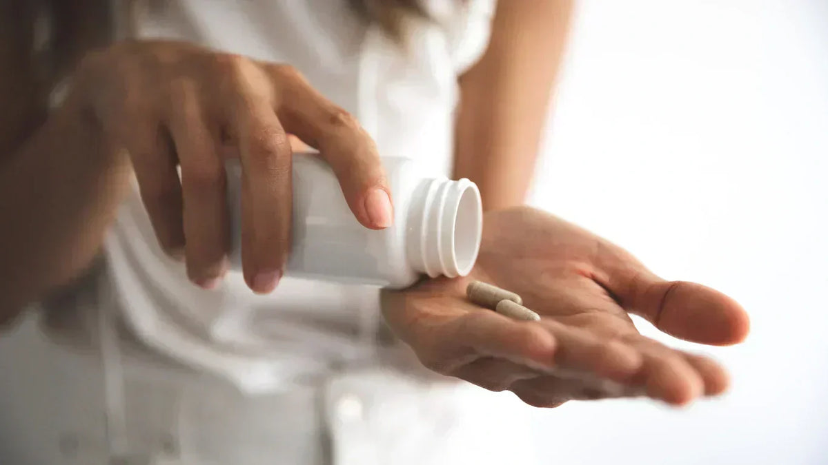 Person tipping supplement capsules from a white bottle into their palm, close-up of hands against a bright background