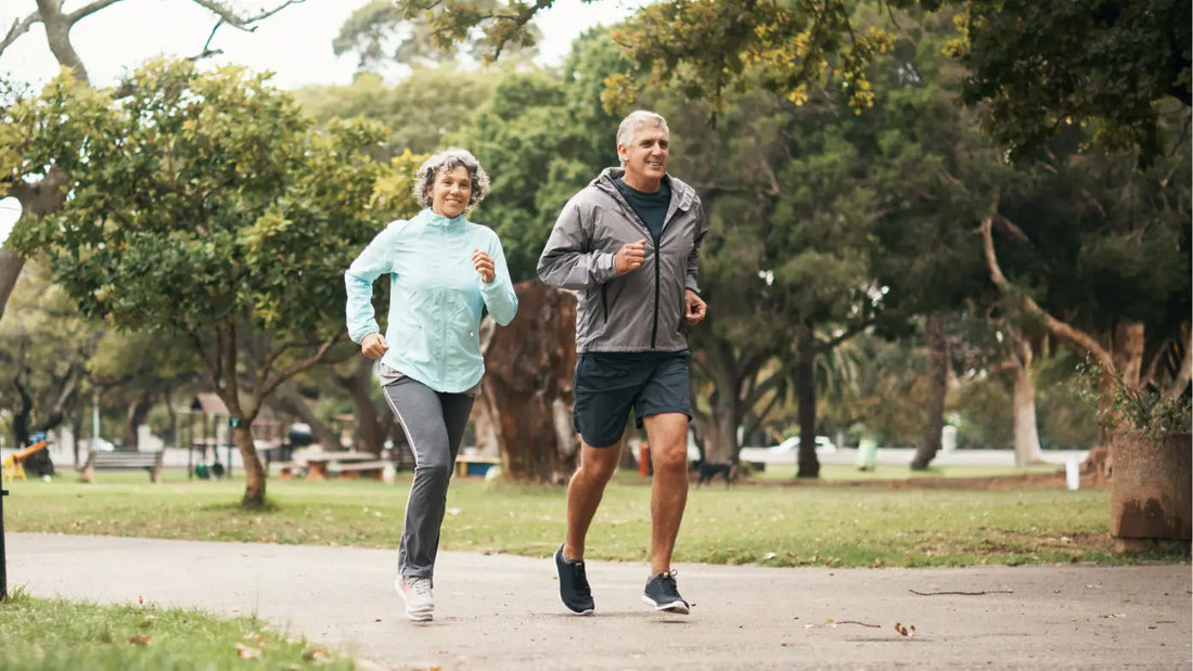 Two older adults – a man and a woman – jogging together on a paved park path surrounded by green trees