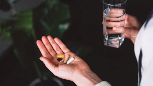 Close-up of a hand holding multiple supplement capsules of different sizes and colours, with a glass of water held in the other hand