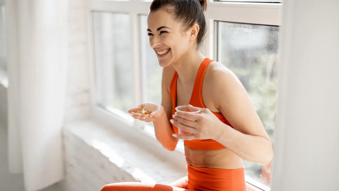 Smiling woman in orange athletic wear sitting by a bright window, holding supplement capsules in one palm and a glass of water in the other hand