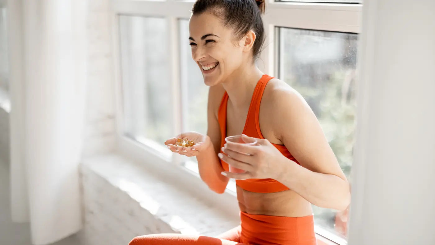 Smiling woman in orange athletic wear sitting by a bright window, holding supplement capsules in one palm and a glass of water in the other hand