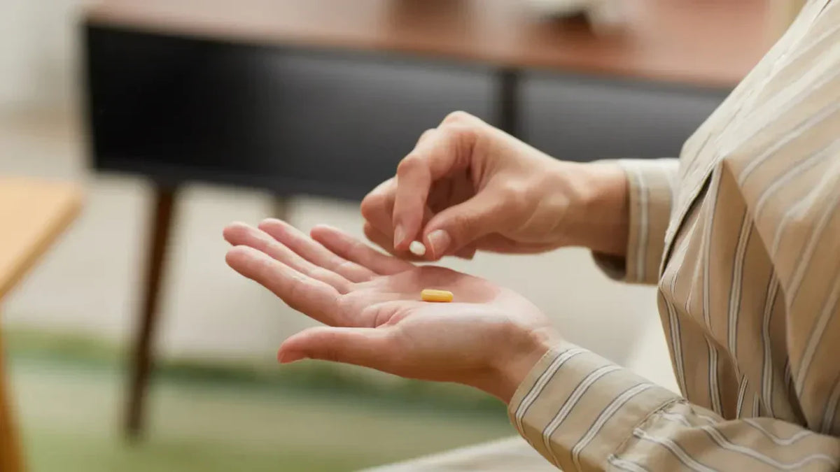Close-up of hands in a striped beige top holding two supplement capsules – one white and one yellow – representing NAD+ and Resveratrol supplements taken together