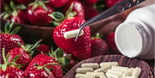 Tweezers holding a supplement capsule over a wooden bowl of capsules set against a background of fresh red strawberries, with a supplement bottle visible