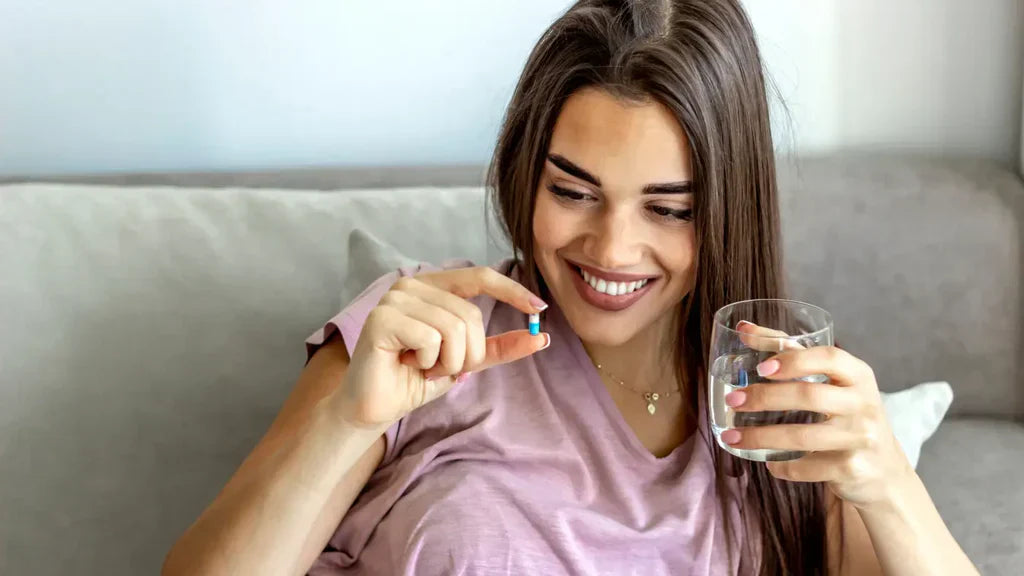 Smiling young woman holding a blue and white supplement capsule and a glass of water while sitting on a grey sofa, illustrating spermidine supplementation