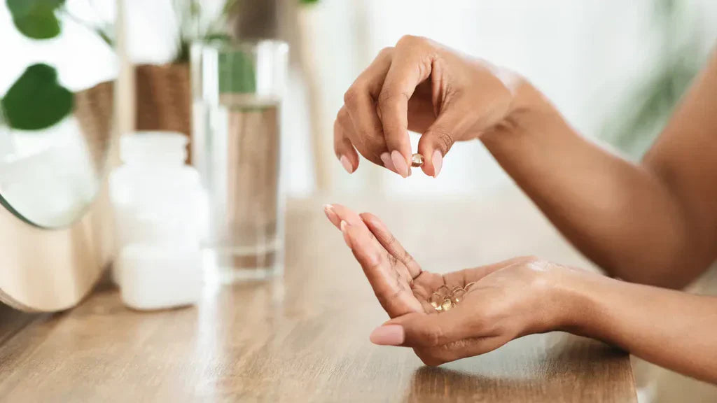 Woman's hands holding a golden supplement softgel over her open palm in a wellness setting with plants and beauty products in the background