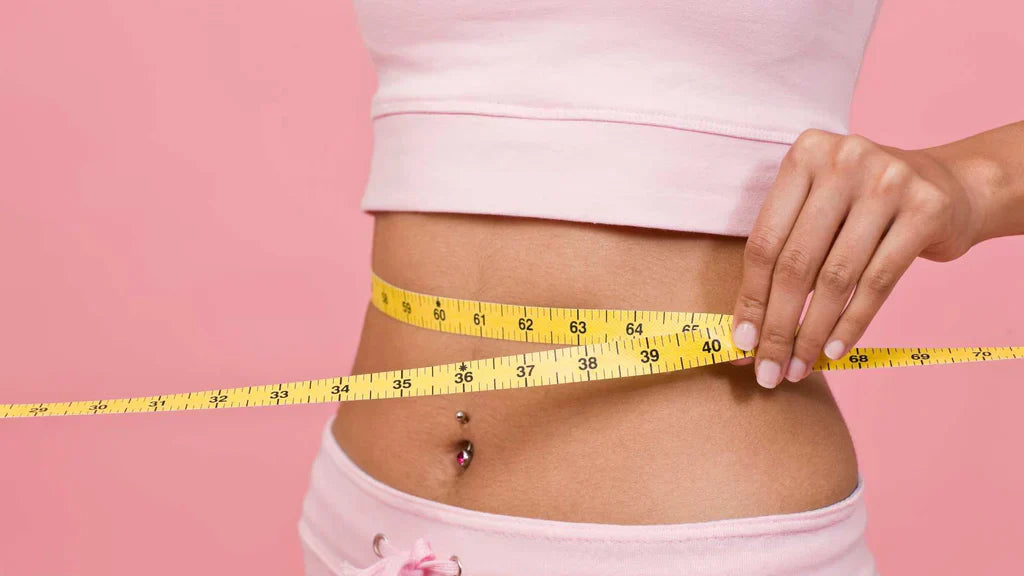 Woman in pink crop top measuring her waist with a yellow tape measure against a pink background, illustrating weight management
