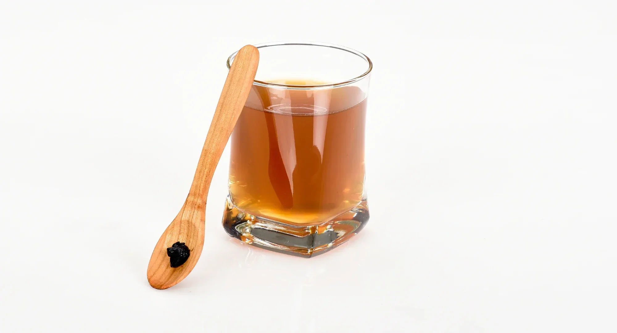 Small glass tumbler containing amber-brown shilajit drink on a white surface, with a wooden spoon holding a piece of shilajit resin resting against the glass