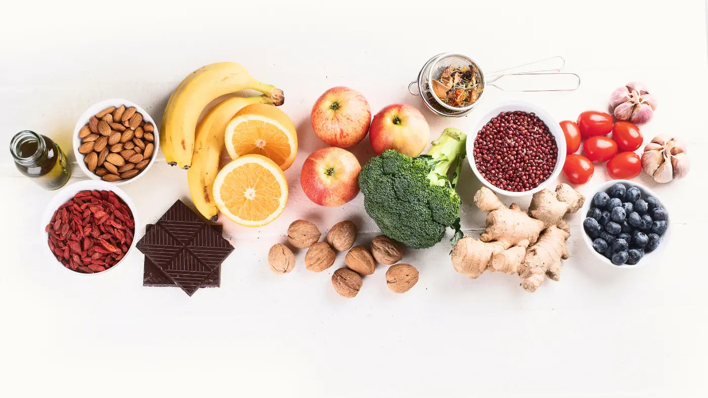 Flat lay of antioxidant-rich foods on a white surface including almonds, banana, orange, apples, broccoli, red berries, tomatoes, garlic, blueberries, walnuts, ginger root, dark chocolate and goji berries