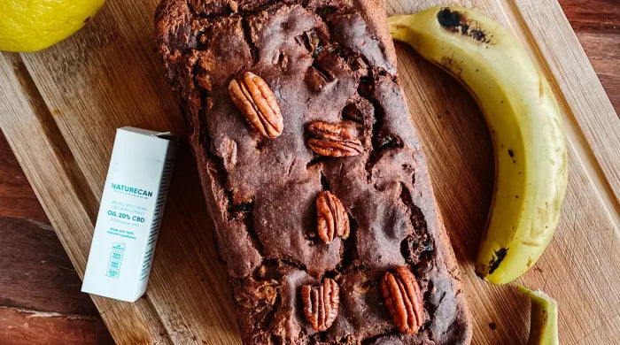 Vegan banana bread loaf topped with pecan nuts on a wooden chopping board, with a Naturecan CBD 20% Oil box, a lemon, and a ripe banana beside it