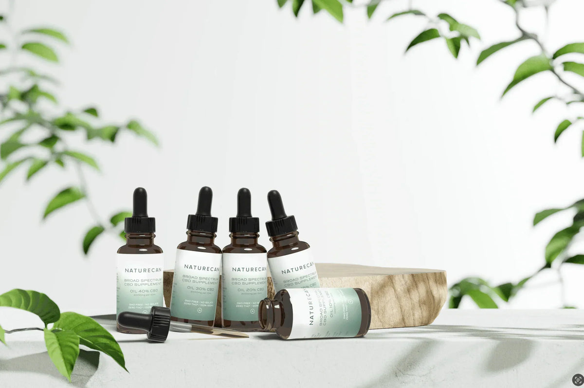Five Naturecan CBD oil dropper bottles arranged on a pale stone surface with lush green plant leaves framing the scene