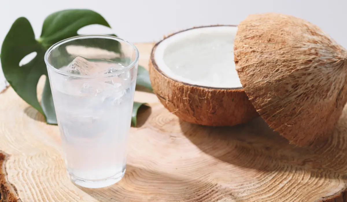 Glass of iced water on a wooden board next to a halved fresh coconut and a green monstera leaf, representing a cortisol cocktail or adrenal cocktail drink