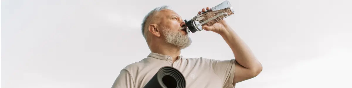 Fit older man with grey beard drinking from a water bottle while carrying a rolled yoga mat outdoors against a pale sky, representing active longevity and healthy ageing