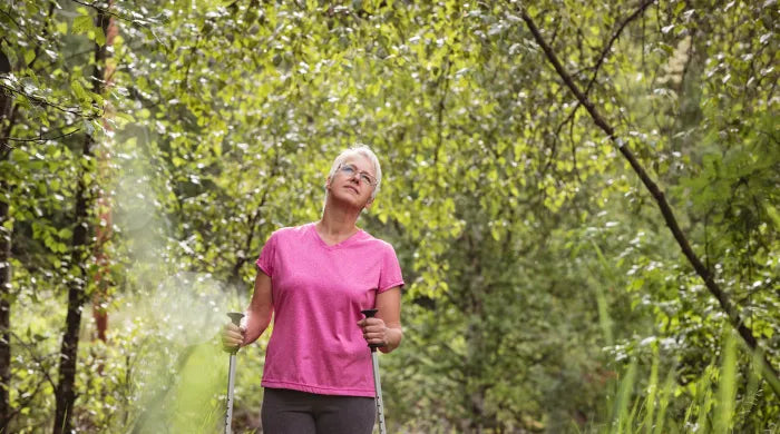 Older woman in a pink t-shirt with glasses nordic walking with poles through a lush green forest trail, representing active wellness during menopause