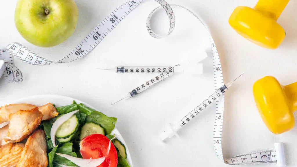 Flat lay of weight management items – a green apple, tape measure, two insulin syringes, a salad bowl with grilled chicken, and yellow dumbbells on a white surface
