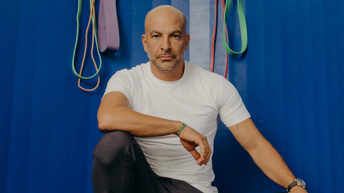 Peter Attia, longevity physician, sitting in a white t-shirt with arms resting on knees in front of a blue wall with colourful resistance bands hanging behind him