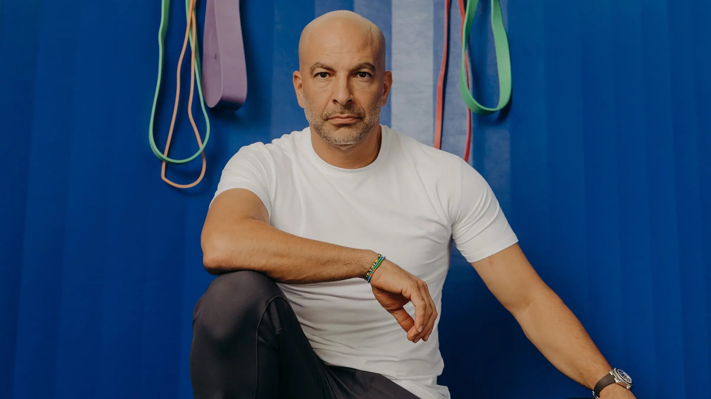 Peter Attia, longevity physician, sitting in a white t-shirt with arms resting on knees in front of a blue wall with colourful resistance bands hanging behind him