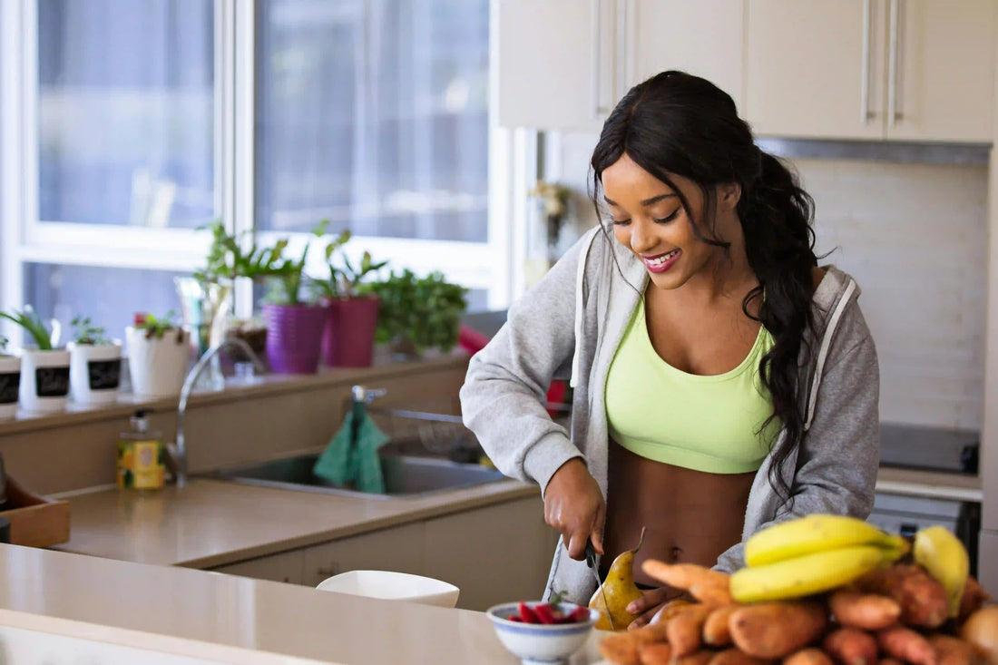Smiling woman in athletic wear chopping fruit and vegetables at a kitchen counter, with potted plants on a bright windowsill in the background