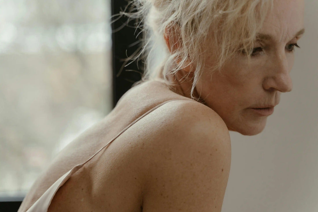 Side profile close-up of an older woman with blonde and white hair and bare shoulder, looking pensive near a window in soft natural light