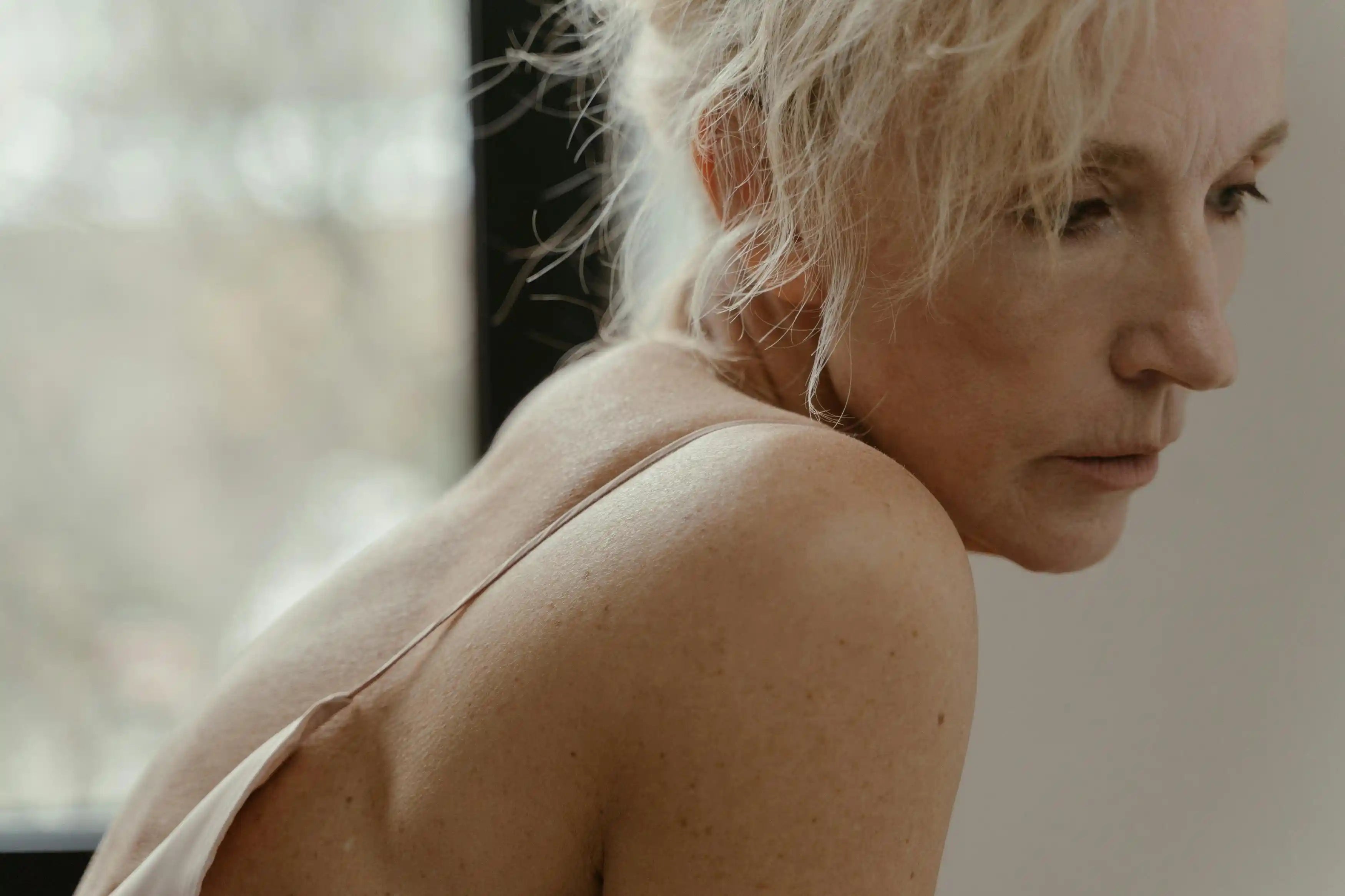 Side profile close-up of an older woman with blonde and white hair and bare shoulder, looking pensive near a window in soft natural light
