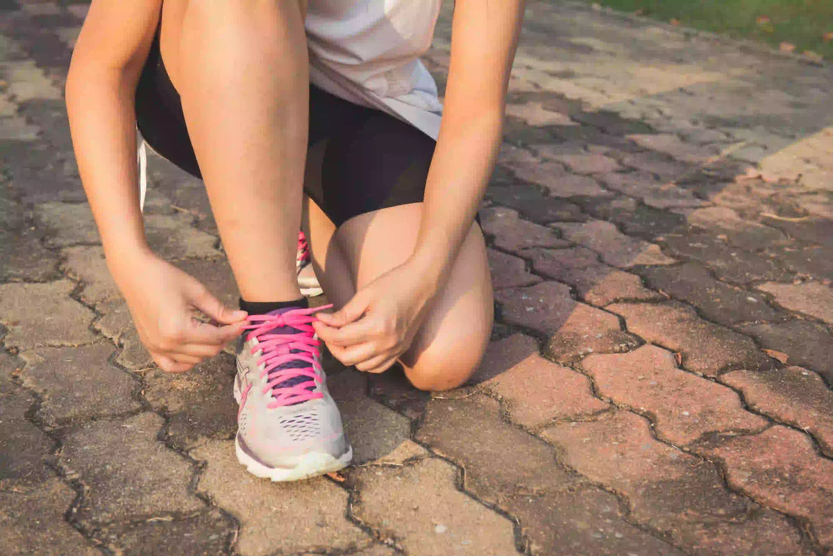Person kneeling down to tie the laces on a pink and grey running shoe on a paved stone surface outdoors