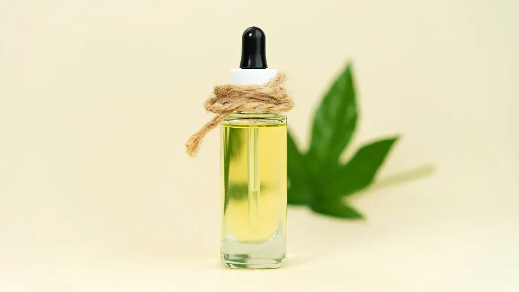 Clear glass dropper bottle containing pale yellow THC oil tied with twine, with a blurred green cannabis leaf in the background on a cream surface