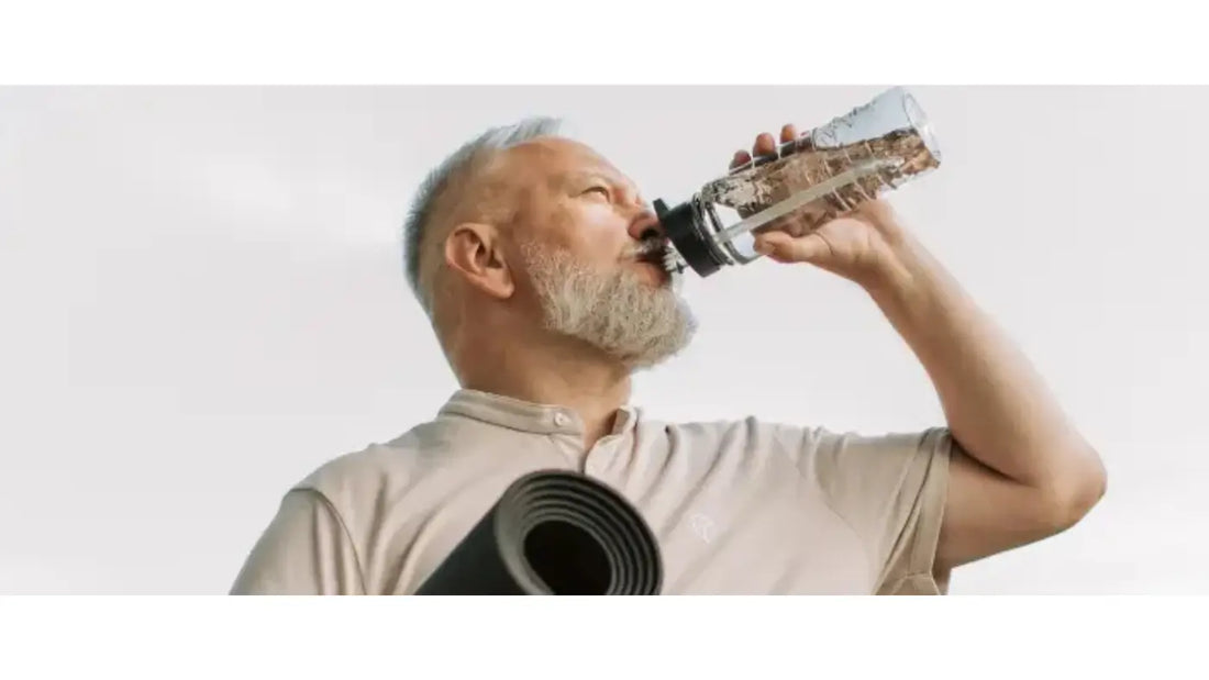 Older man with grey hair and beard drinking from a clear water bottle while carrying a rolled yoga mat, photographed against a light background