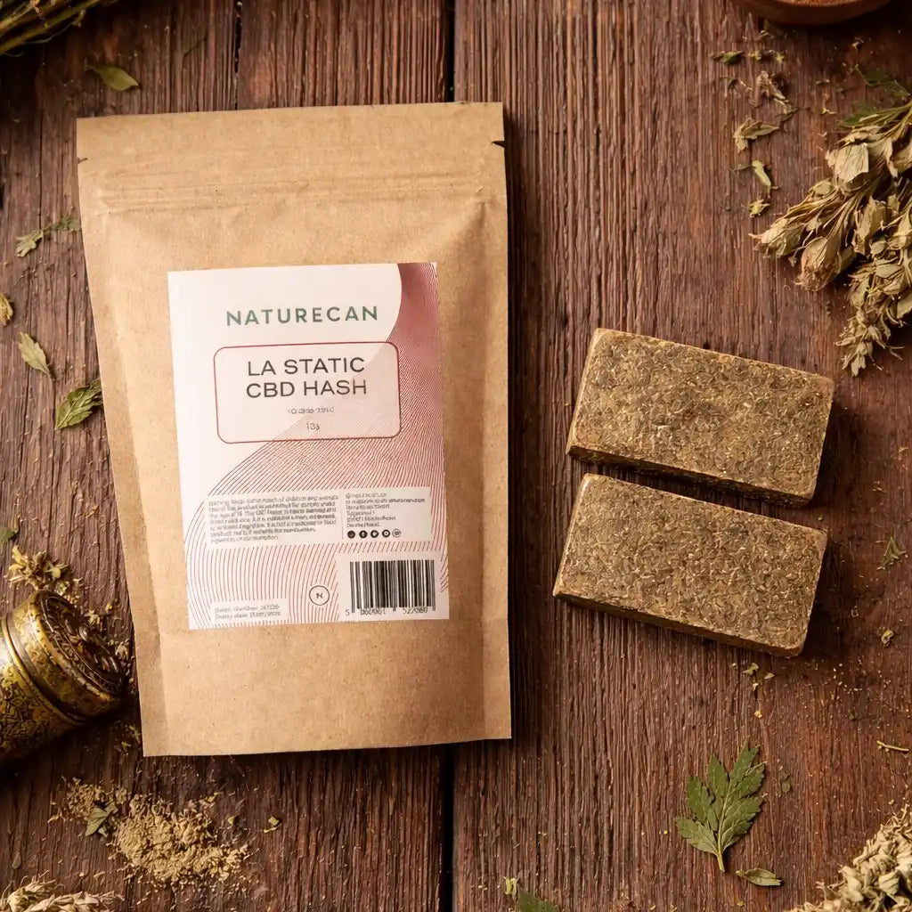 Naturecan LA Static CBD Hash lifestyle – kraft paper pouch with pink and coral label on dark rustic wooden surface surrounded by dried hemp leaves and mortar, with hash blocks beside it