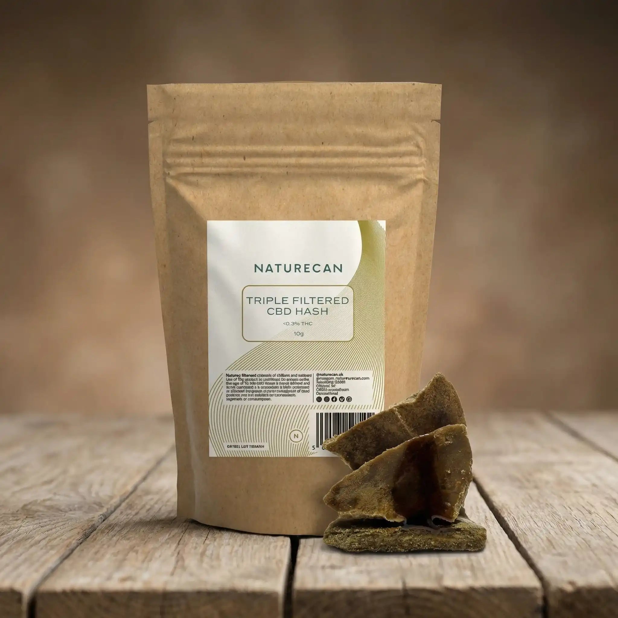 Naturecan Triple Filtered CBD Hash lifestyle – kraft paper pouch with cream and gold label, Triple Filtered CBD Hash less than 0.3% THC 10g, on rustic wooden surface with broken chunks of hash beside it