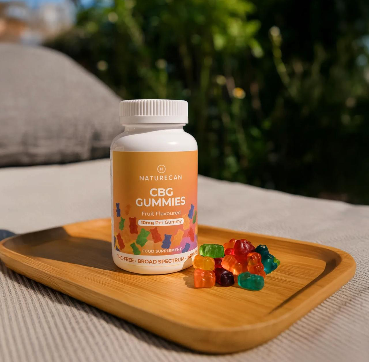 Naturecan CBG Gummies lifestyle – bottle on wooden tray in outdoor garden setting, Fruit Flavoured, 10mg per gummy, THC-Free, Broad Spectrum, with colourful gummy bears scattered around it